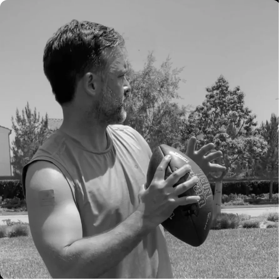 A black and white photo of a man in a sleeveless shirt holding an American football outdoors.