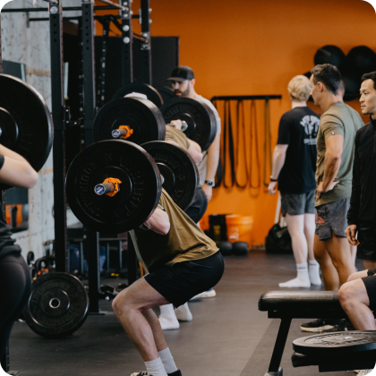 A person performs a barbell squat in a gym while others watch in the background.
