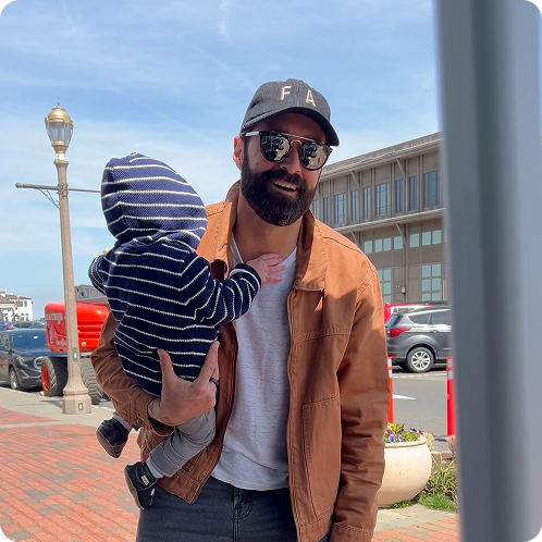 A man with a beard and sunglasses holds a baby with its back to the camera on a sidewalk.