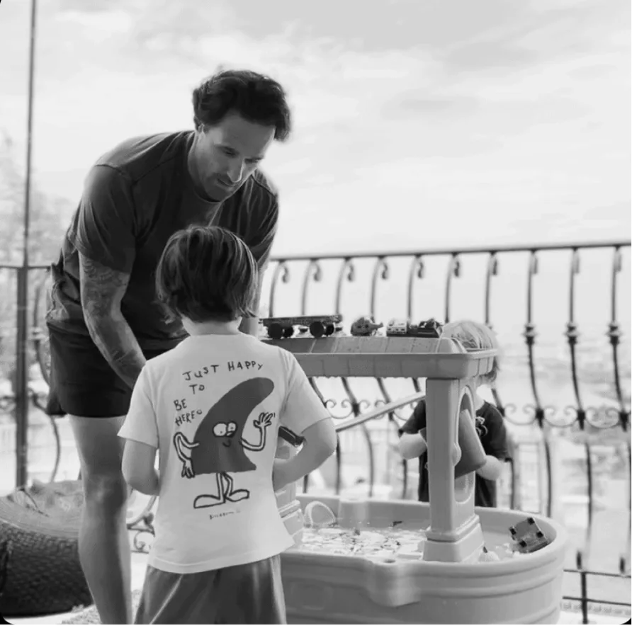 A black and white photo of a father playing with his two children at a water table on a balcony.