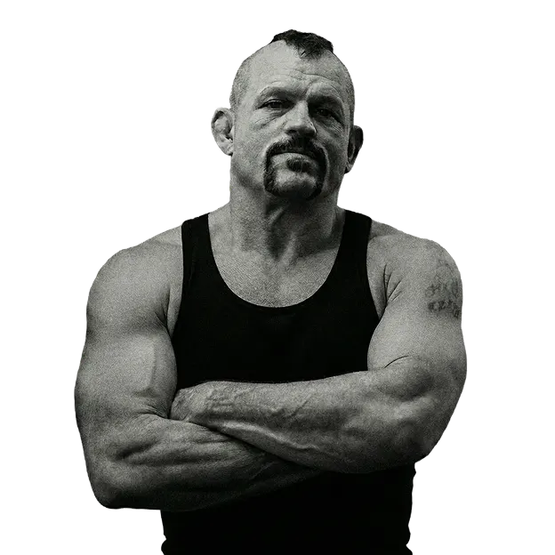 A muscular man with a mohawk and mustache stands with his arms crossed in a black and white photo.