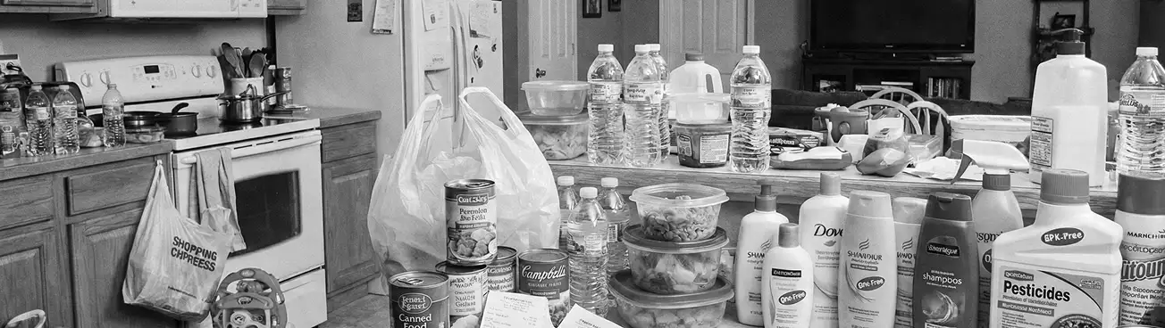A black and white photograph of a kitchen with counters covered in groceries, water bottles, and other supplies.