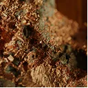 A close-up photograph of raw, coppery mineral flakes mixed with dark rock.