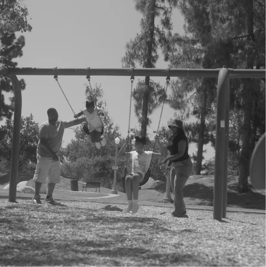 A black and white photo of two adults pushing two children on a swing set at a park.