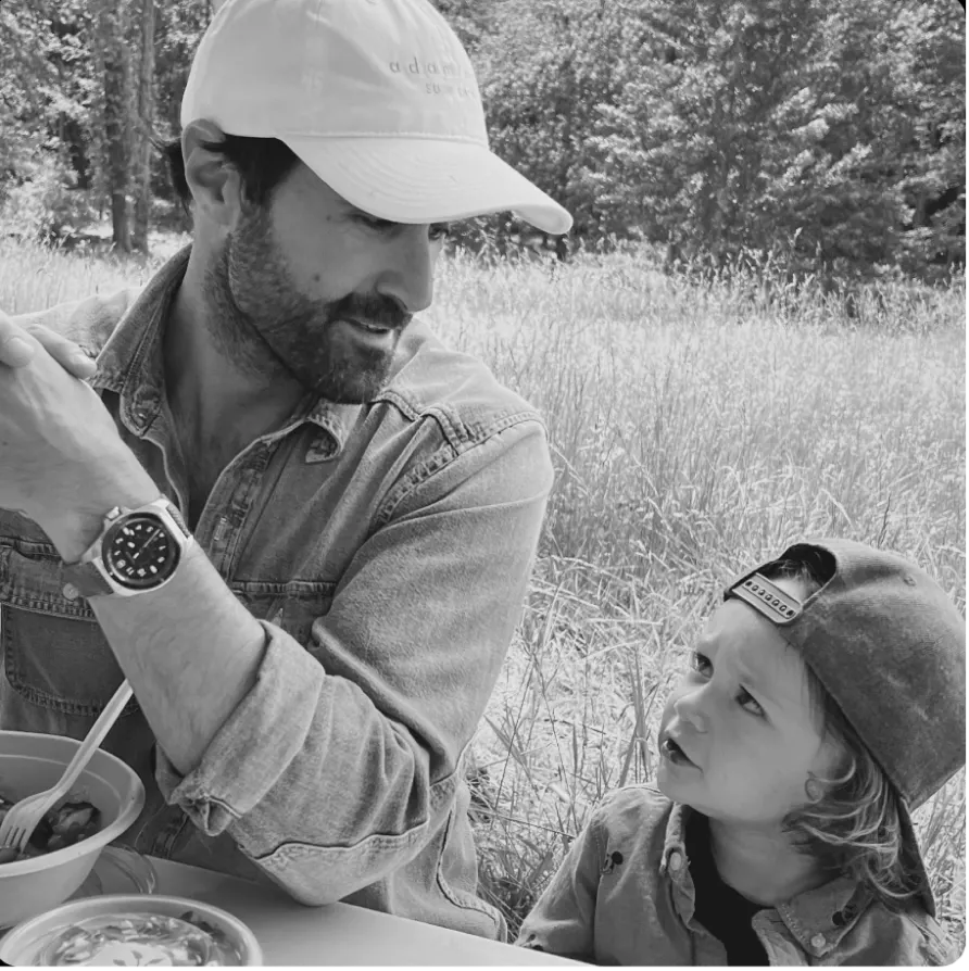 A black and white photo of a man and a young child in hats talking while having a meal outdoors.