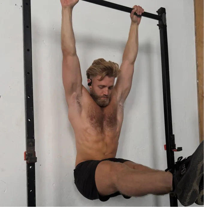 A shirtless man with blond hair doing a hanging leg raise on a pull-up bar.