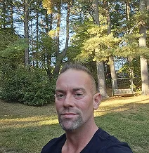 Person sitting outdoors in a wooded area with sunlight filtering through the trees.