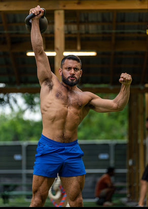 A shirtless, muscular man lifts a black kettlebell overhead with one arm while outdoors.