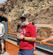 Man in red shirt and cap standing on a train, with rocky landscape in background.