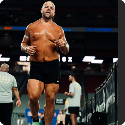 A sweaty, shirtless man wearing a backwards cap and black shorts runs during an indoor competition.