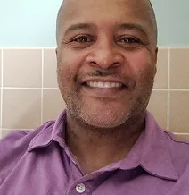 Smiling man in a purple shirt, in front of a tiled wall.