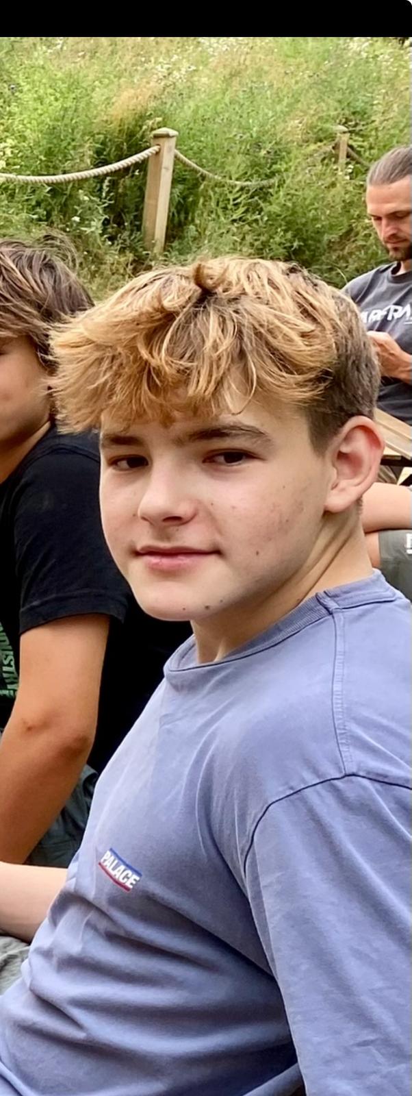Young person with light hair outdoors, sitting with others.