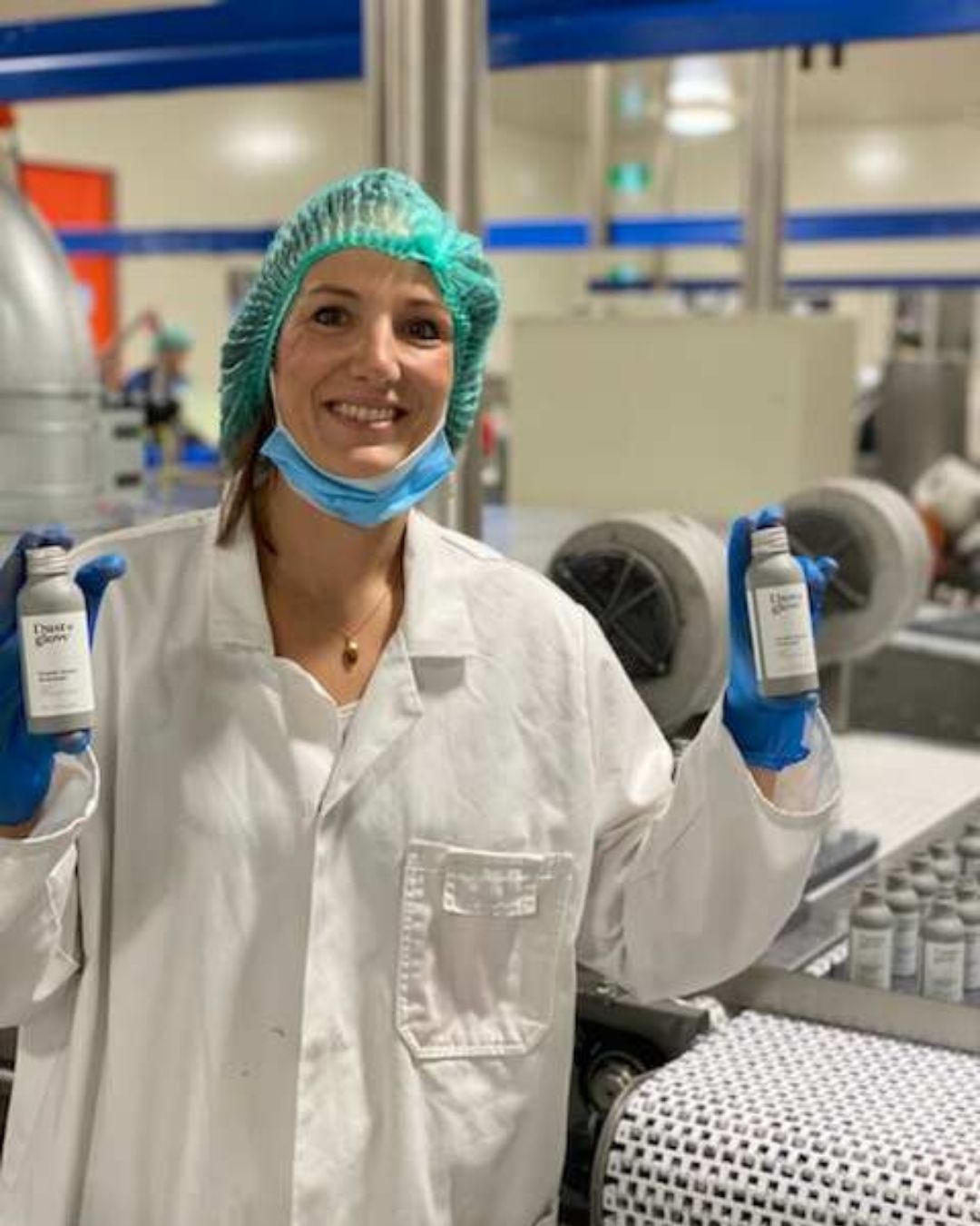 Person in lab coat and hairnet holding two bottles in a factory setting.