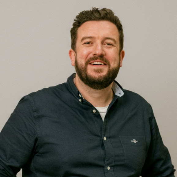 A smiling man with a beard, wearing a dark blue button-down shirt against a plain background.