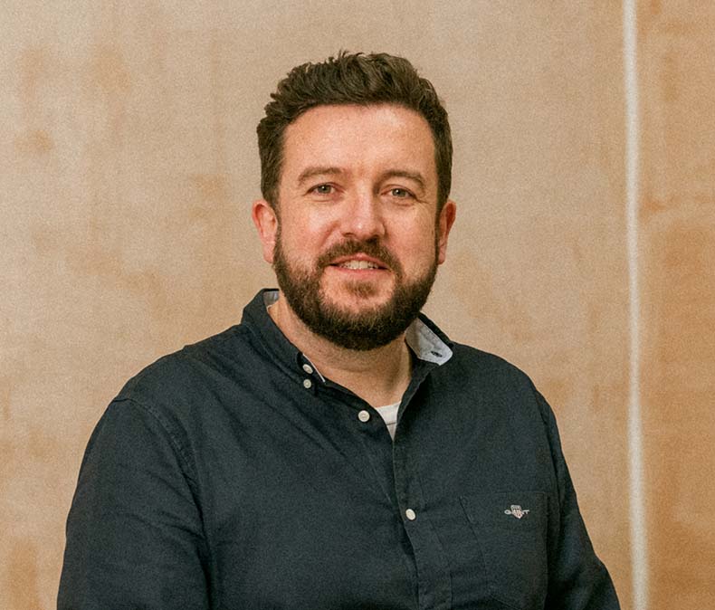 A man with brown hair and a beard, wearing a navy button-down shirt, smiles at the camera.