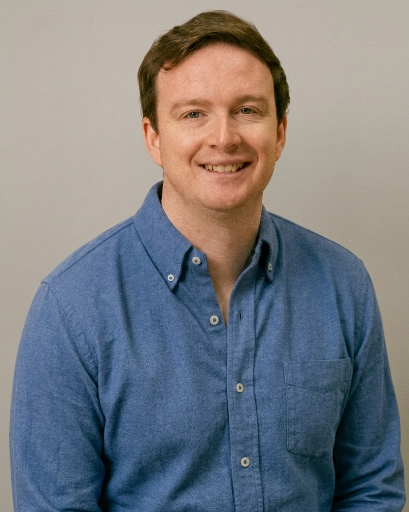 A smiling man with brown hair wearing a blue button-down shirt, photographed from the chest up.