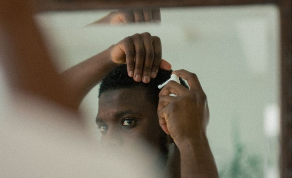 A man's reflection in a mirror as he sprays a product into his hair with one hand.
