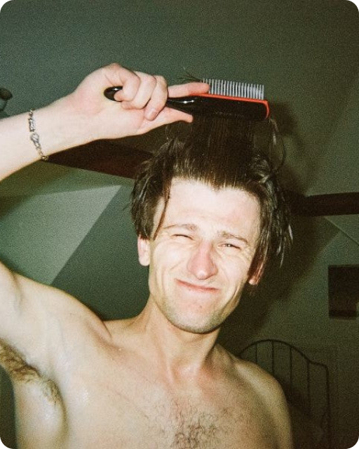 A shirtless man scrunches his face in a smile while brushing his wet hair.