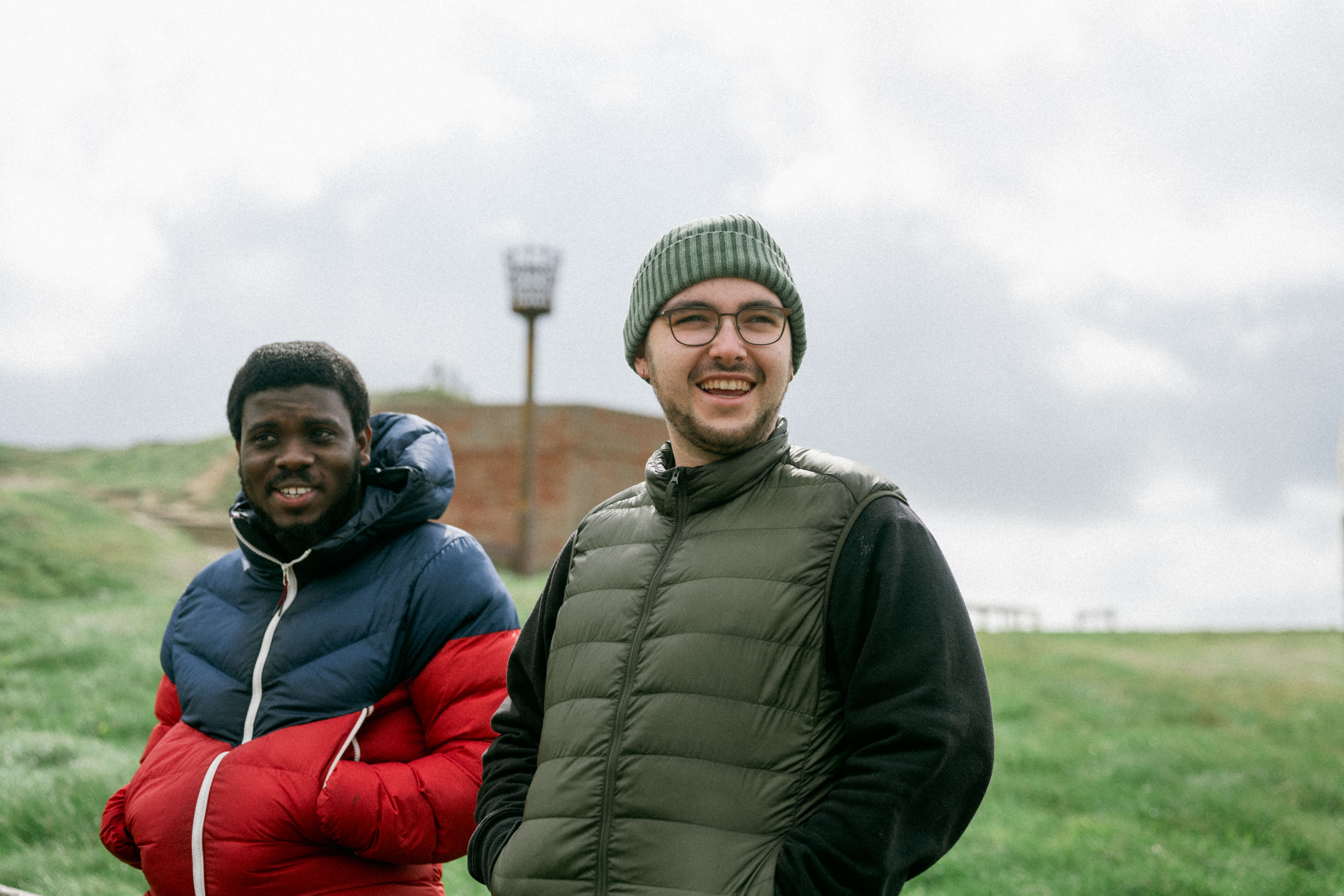 Two men in warm jackets standing together in a grassy field on a cloudy day.