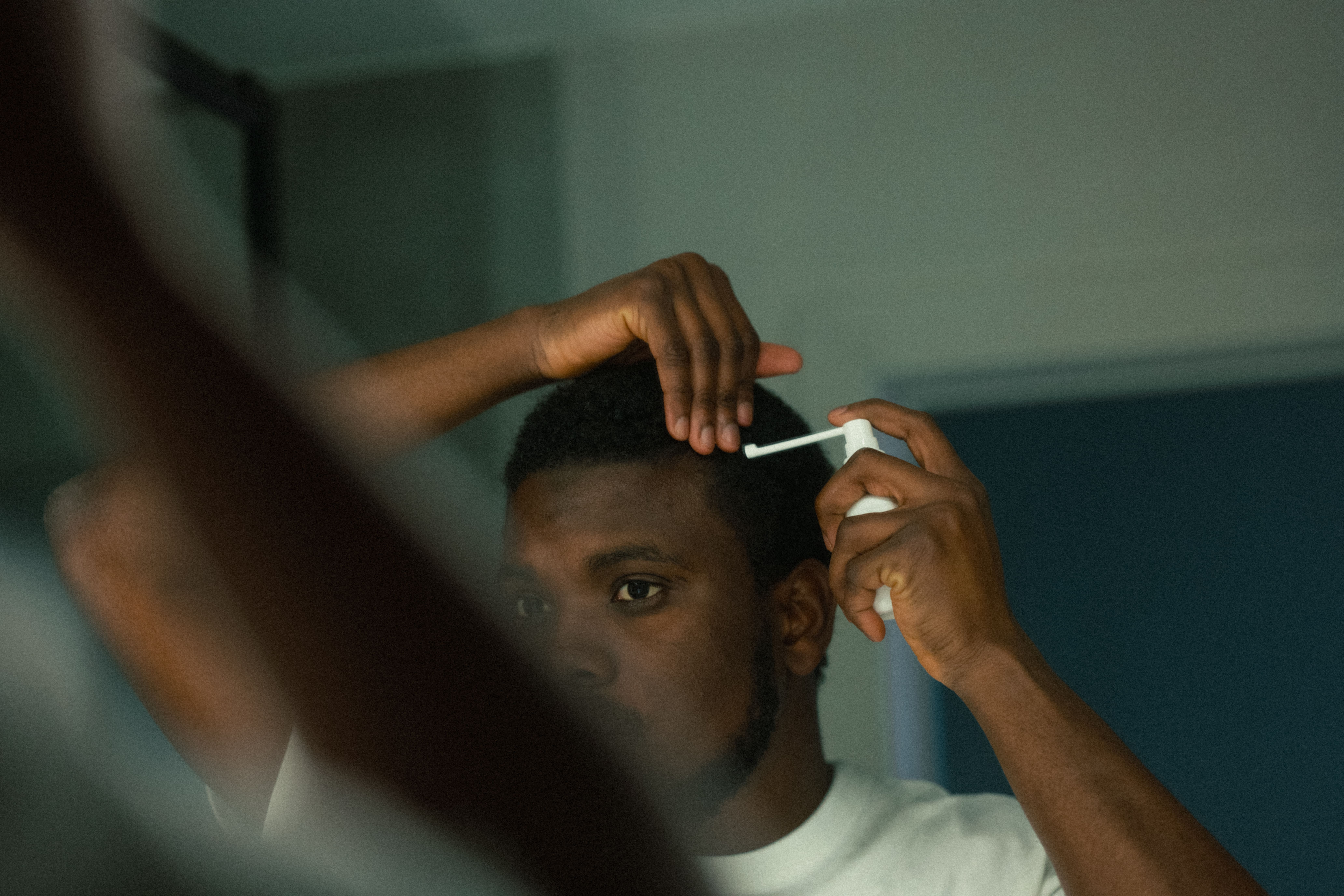 A man looks in a mirror while applying a spray product with a nozzle to his hair.