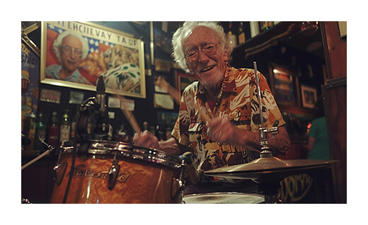 Elderly man playing drums in a dimly lit room with framed pictures.