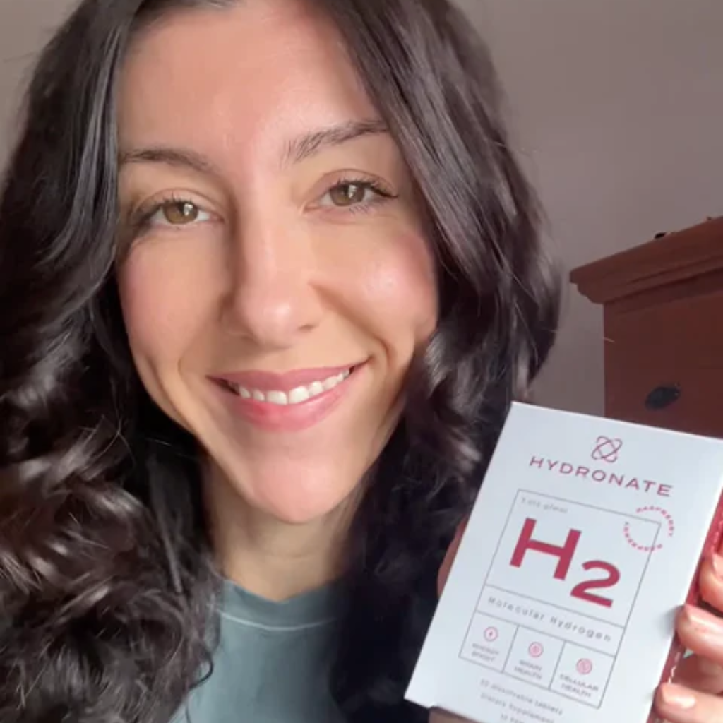 A smiling woman with dark, wavy hair holds up a white box of Hydronate H2 supplement.