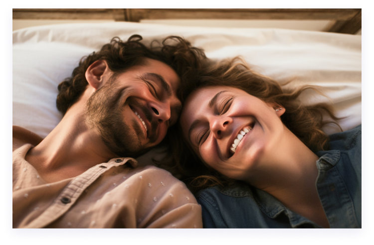 An overhead shot of a man and woman lying side-by-side in bed, smiling with their eyes closed.