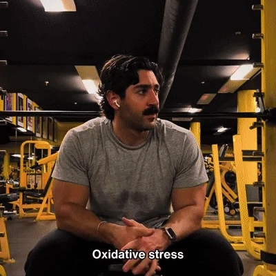 A muscular man with a mustache wearing a grey t-shirt and earbuds sits in a gym.