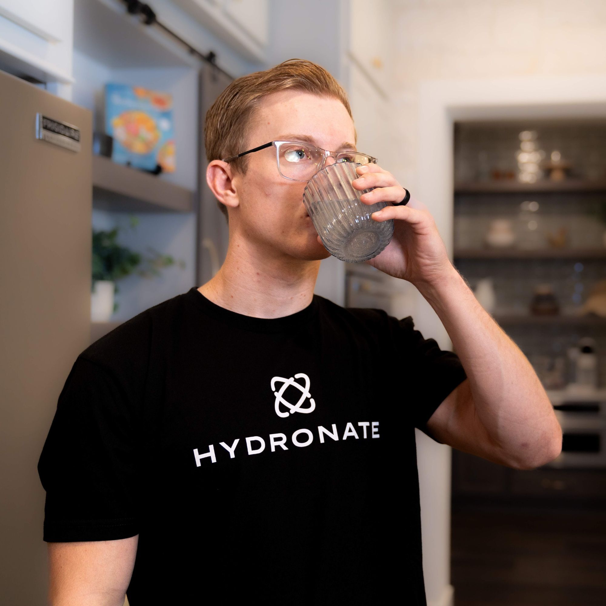 Person wearing a 'Hydronate' t-shirt drinking from a clear glass in a kitchen.
