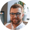 A circular headshot of a smiling man with a beard and glasses.