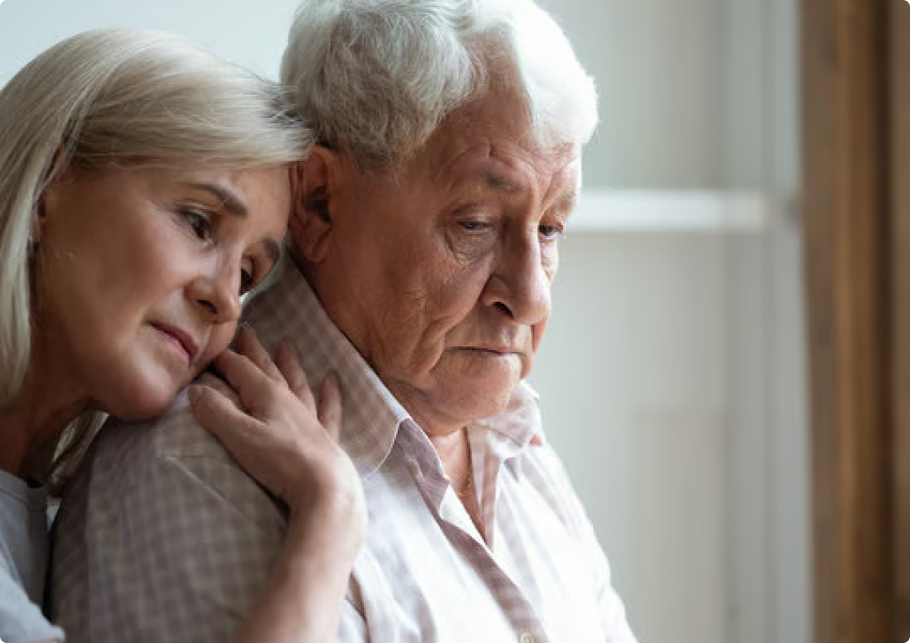 An elderly woman comforts a sad-looking elderly man by resting her head and hand on his shoulder.