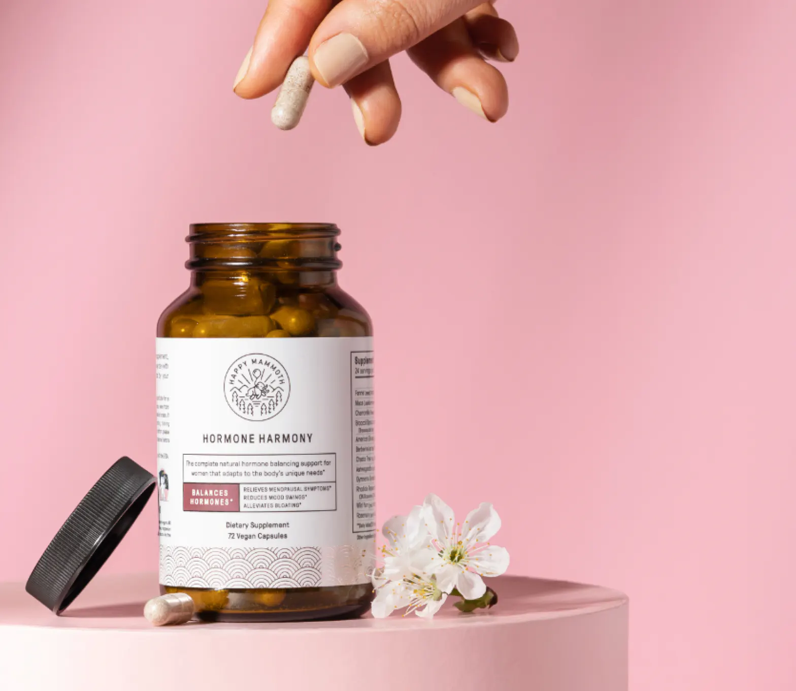 Hand holding capsule over open supplement bottle beside flowers.
