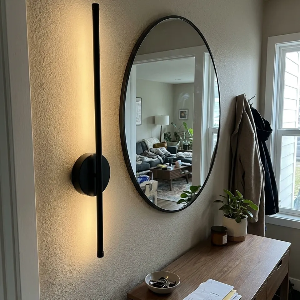 A modern black vertical wall sconce hangs on a wall next to an oval mirror in an entryway.