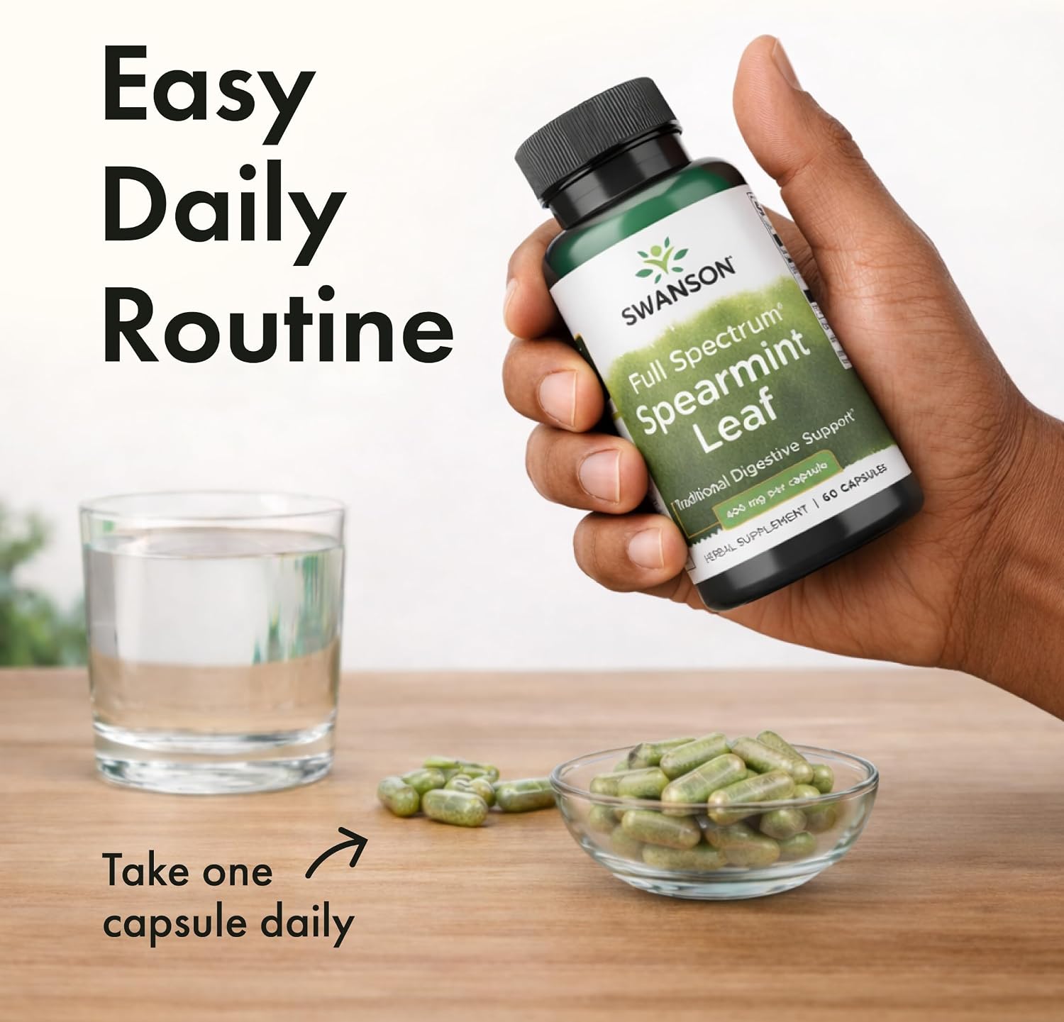 A hand holds a bottle of Swanson Spearmint Leaf supplements over capsules in a bowl and a glass of water.