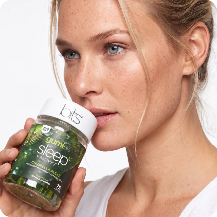 A woman with freckles holds a bottle of gumi bits sleep + recovery supplements near her face.
