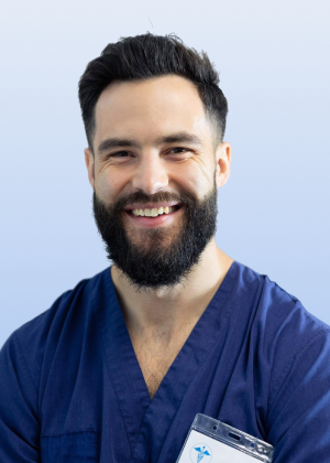 A smiling man with a dark beard wearing blue medical scrubs and an ID badge.