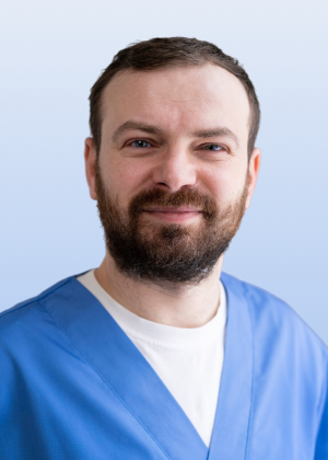 A headshot of a smiling man with a beard, wearing blue scrubs against a light blue background.