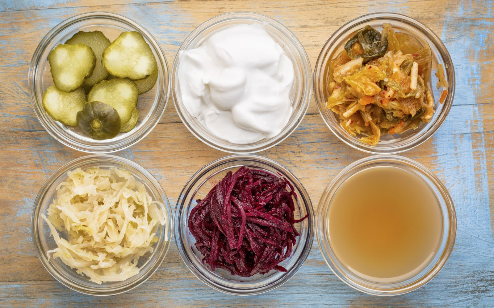 An overhead shot of six small glass bowls containing various fermented foods on a light blue wooden surface.