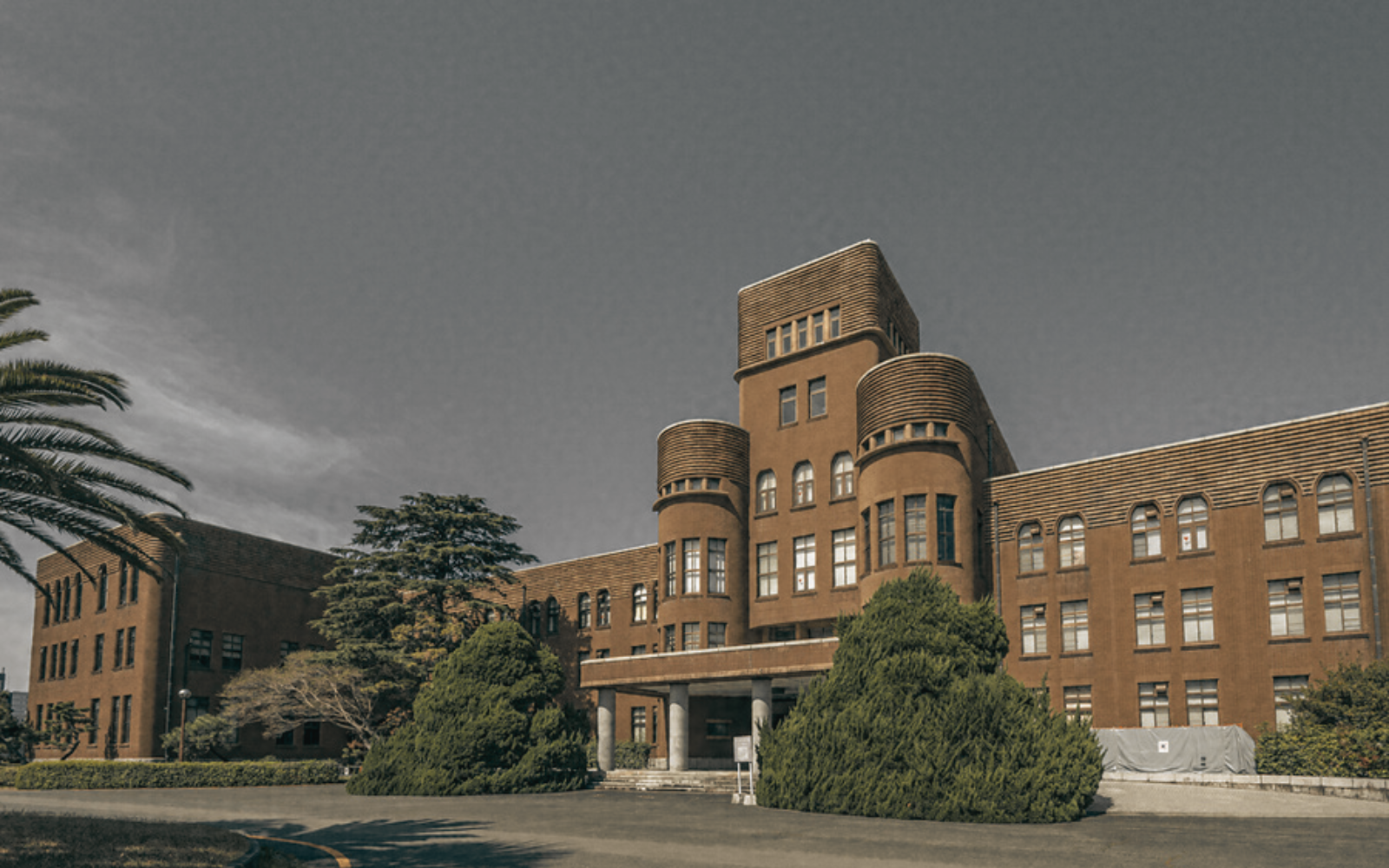 A large, brown institutional building with rounded towers and arched windows, surrounded by trees under a grey sky.