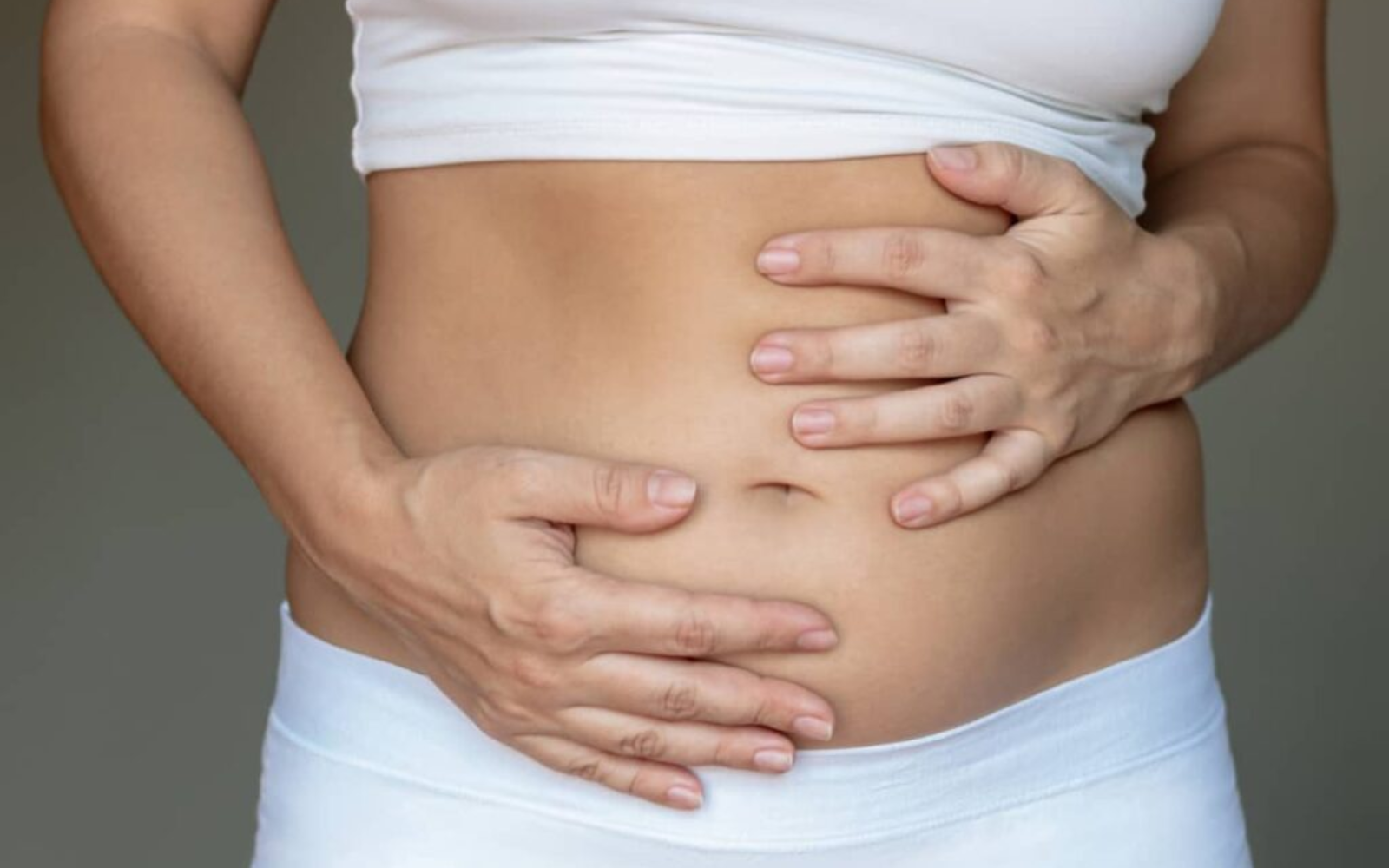 A close-up of a person in white undergarments holding their stomach with both hands against a grey background.
