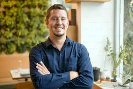 Person smiling with arms crossed in a room with plants and a window.
