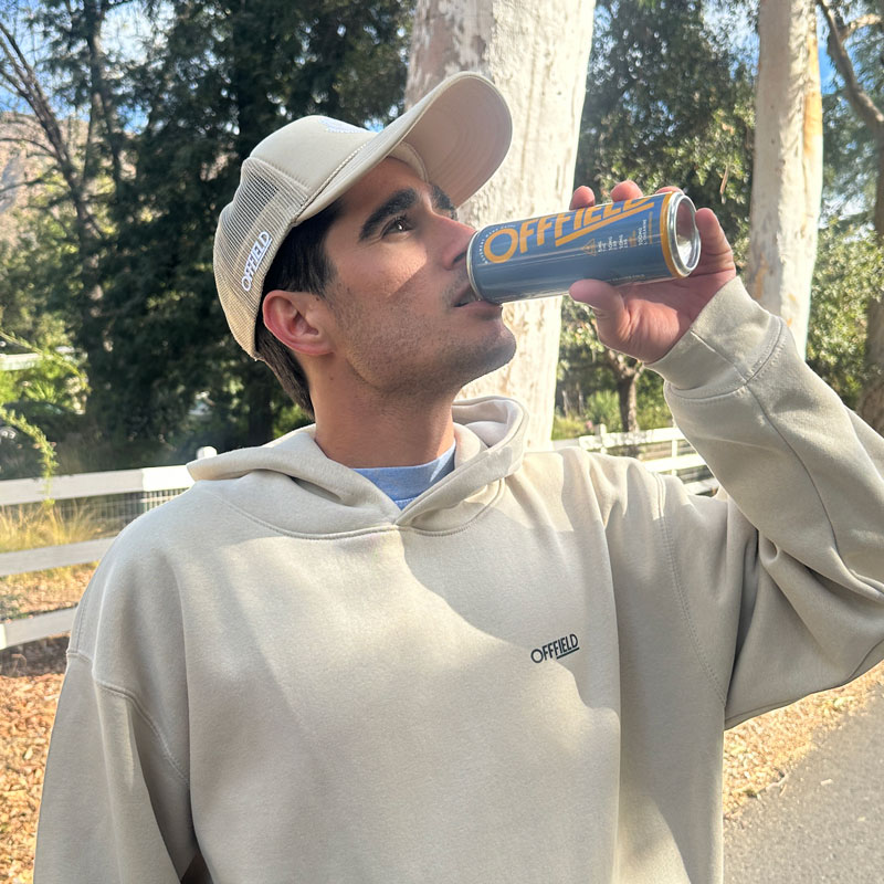 Person in a beige hoodie and cap drinking from a can outdoors.