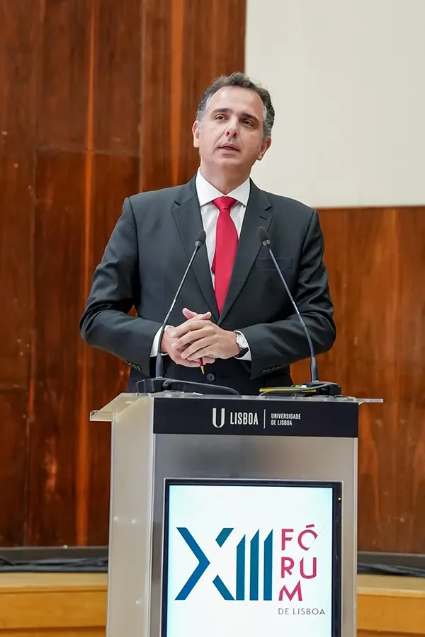 A man in a black suit and red tie stands behind a podium speaking into microphones.