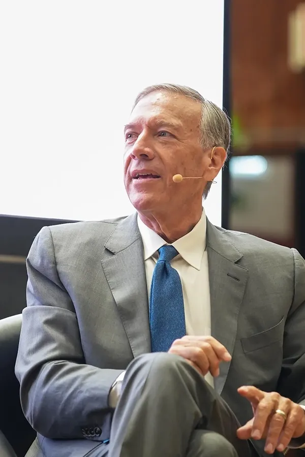 A man in a grey suit, white shirt, and blue tie, wearing a headset microphone while sitting.