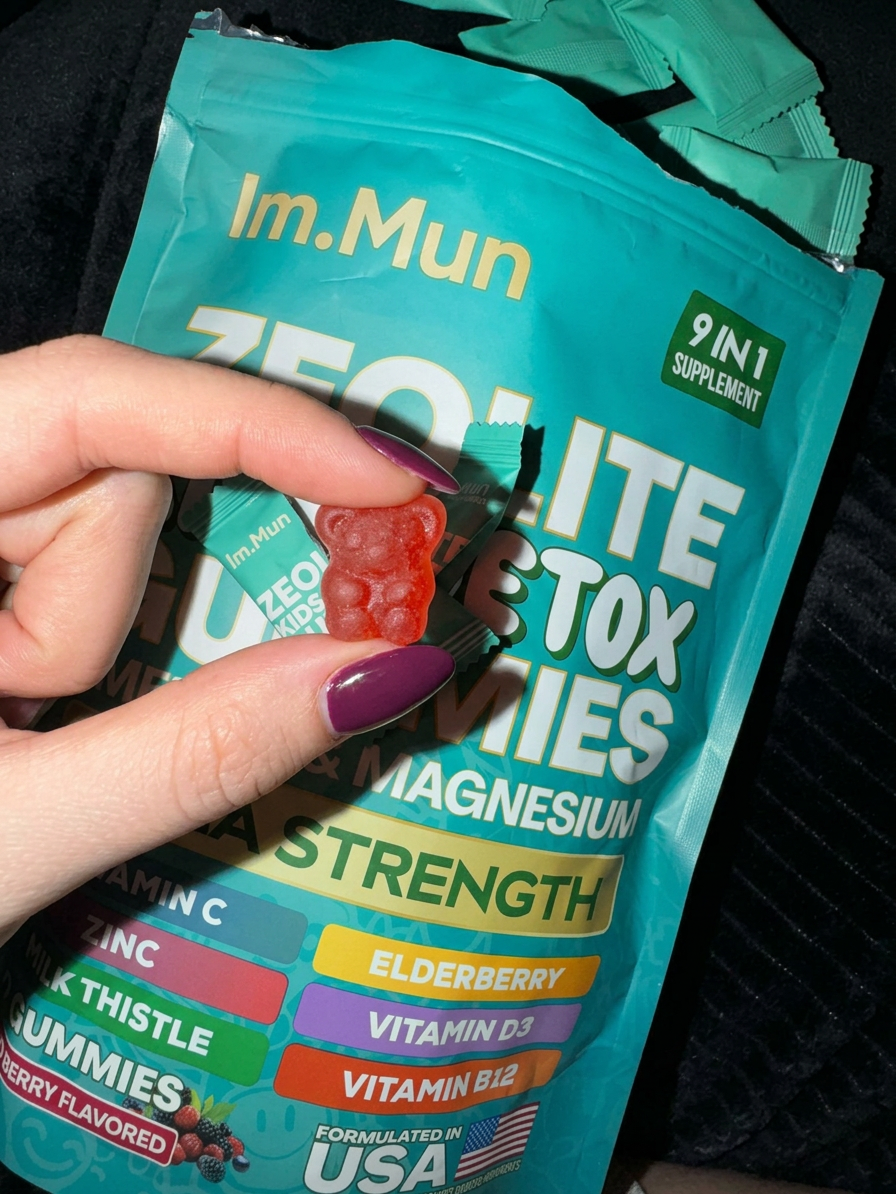 A hand with dark red nail polish holds a red, bear-shaped gummy in front of a supplement pouch.