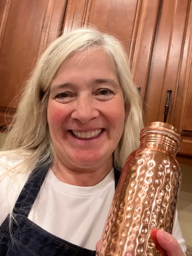 A smiling woman with blonde hair holds up a hammered copper water bottle in a kitchen.
