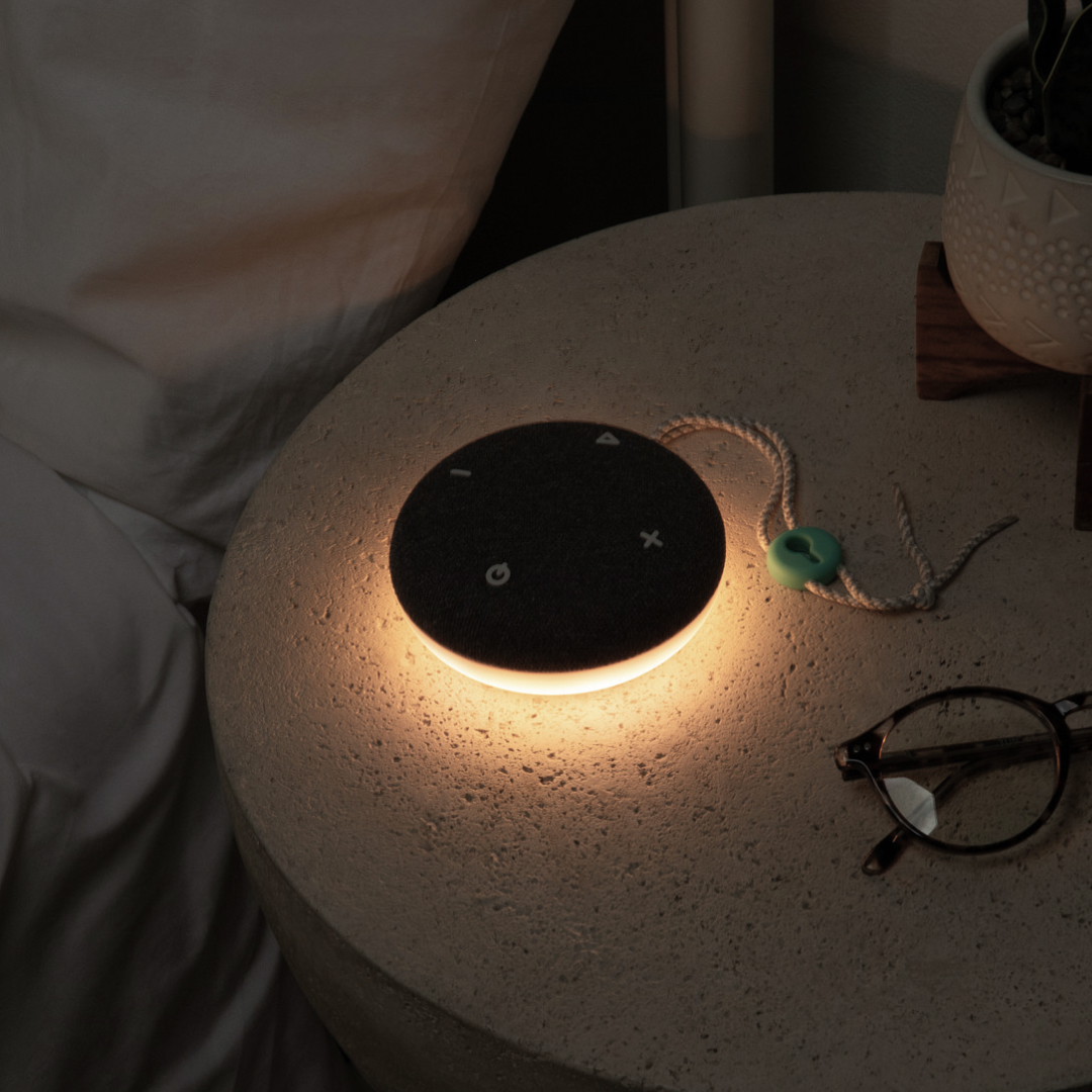 An illuminated SNOOZ Go device  rests on a nightstand next to a pair of eyeglasses.