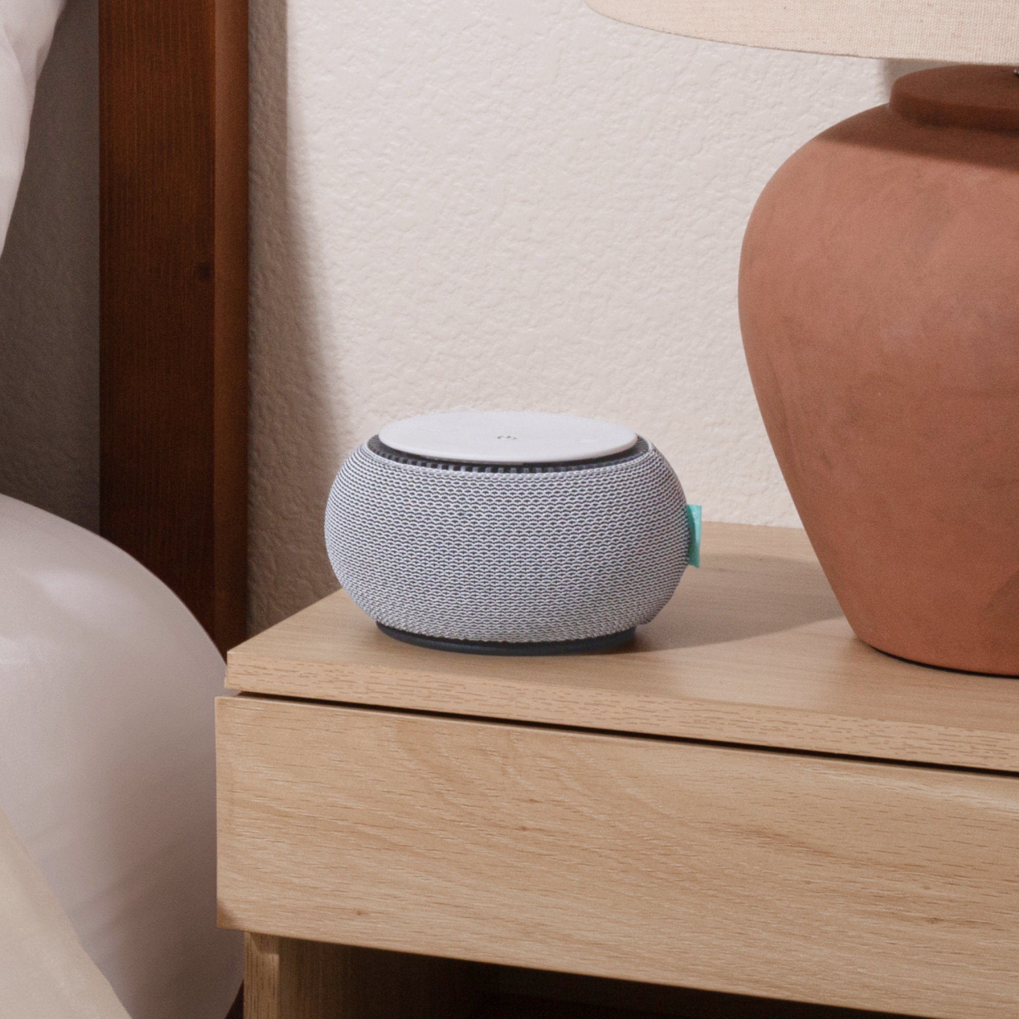 A small, grey and white sound machine sits on a light wood nightstand next to a terracotta lamp.
