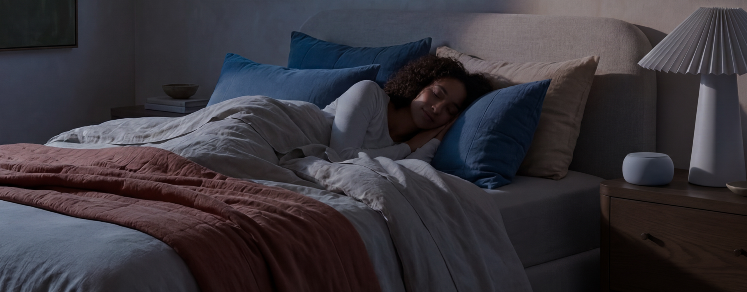 A woman sleeps peacefully in a bed in a dimly lit room, next to a nightstand.