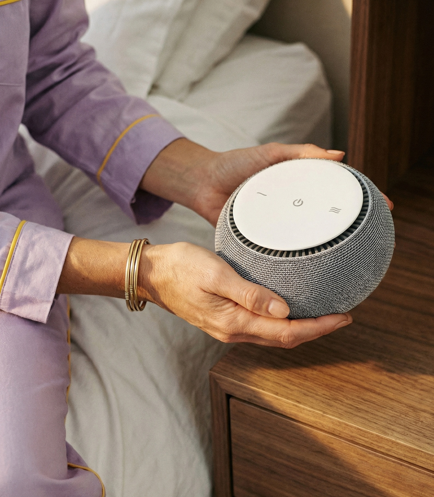 A person in purple pajamas holds a round, grey electronic device over a wooden nightstand.